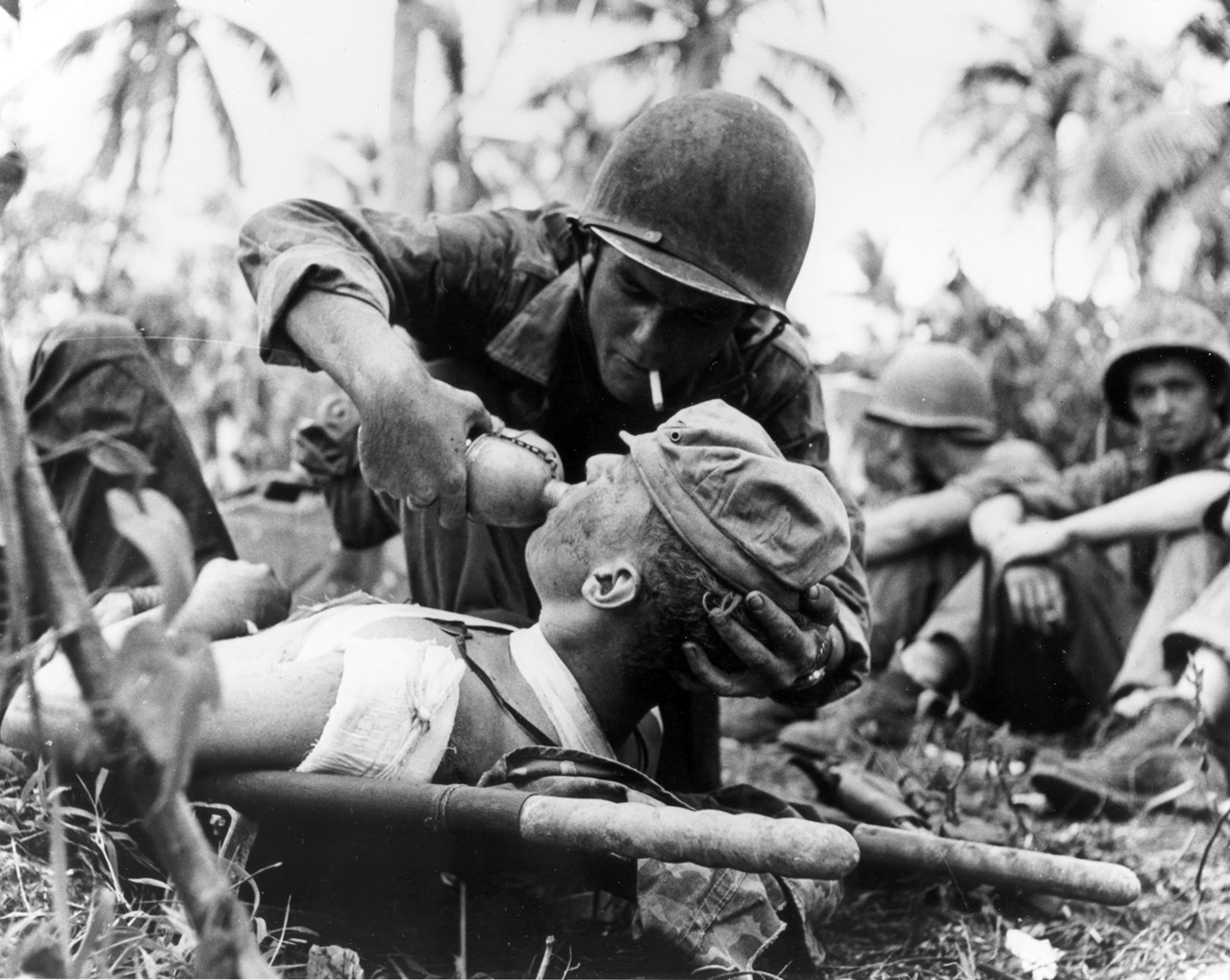 A soldier with a cigarette tucked in his mouth gives a wounded solider lying on a stretcher a drink of water from a canteen.