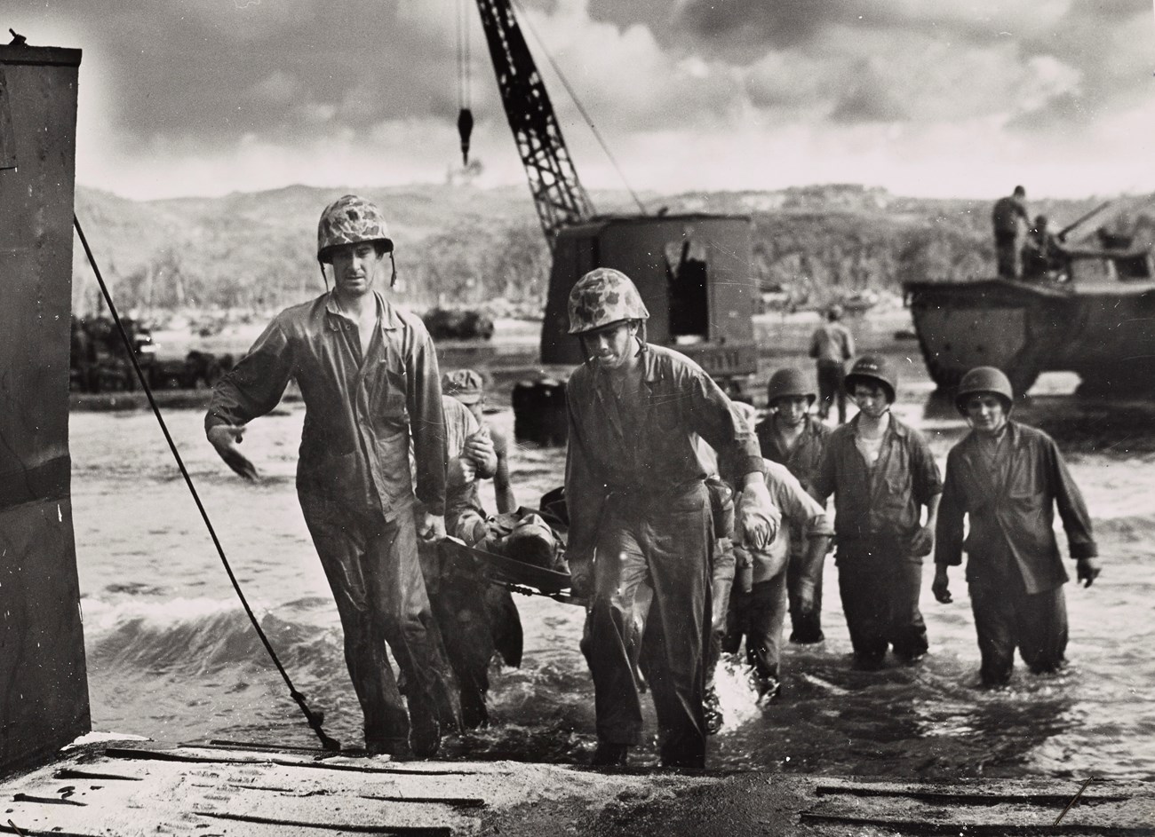 Uniformed soldiers carry a wounded man on a litter through the water to a hospital ship.