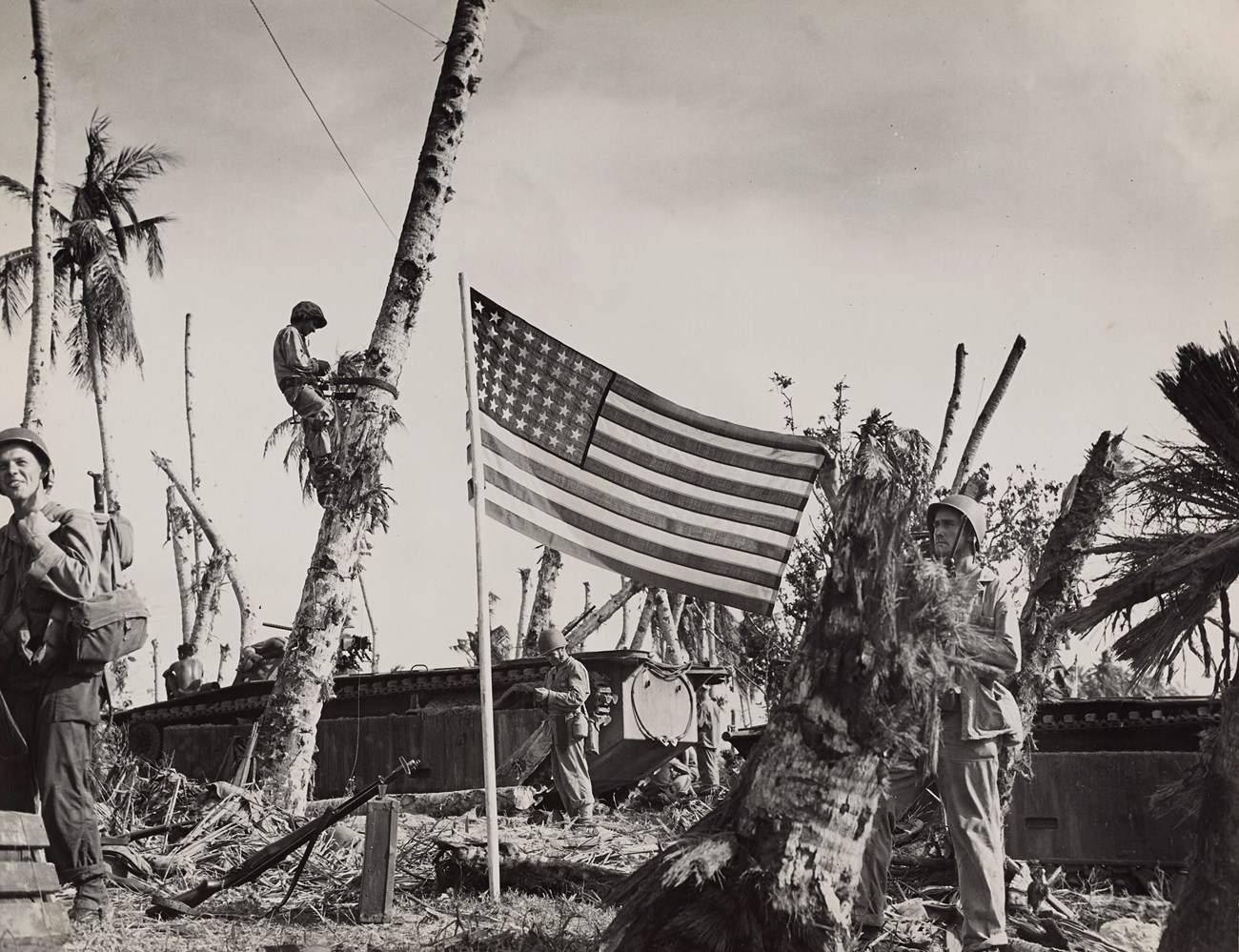 An American flag flies from a pole on a beach. Four armed soldiers are working and two amtracs are behind the flag. The area has been severely bombed.