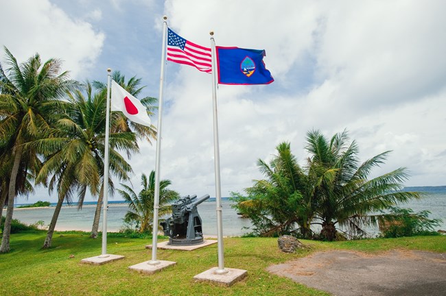 Three flag poles flying the Japanese, American and Guam flag in front of a large mounted coastal defense gun overlooking the ocean.