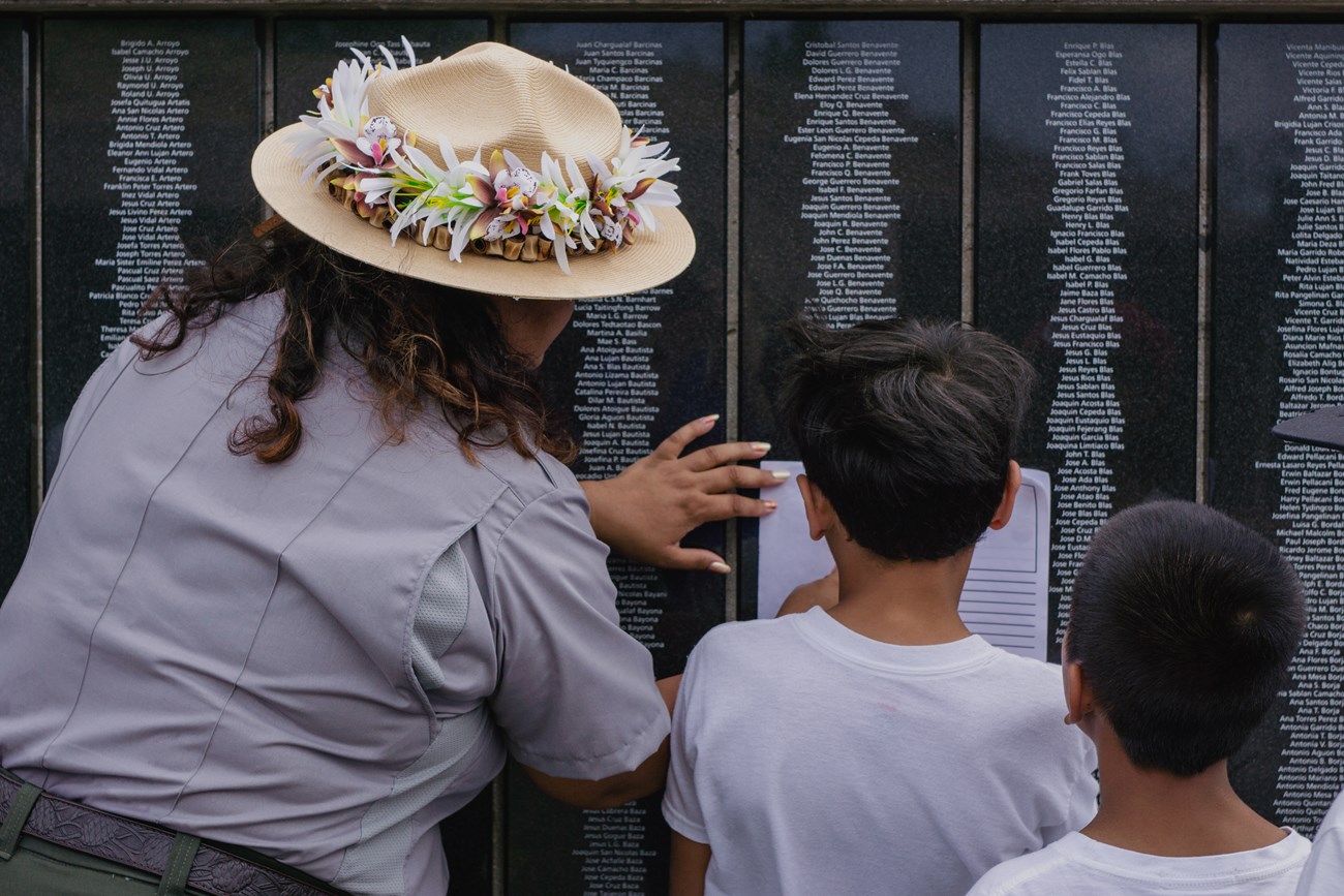 A woman in a park ranger uniform with flower around the brim of her hat helps a group of children find names on a black plaque covered with engraved names.