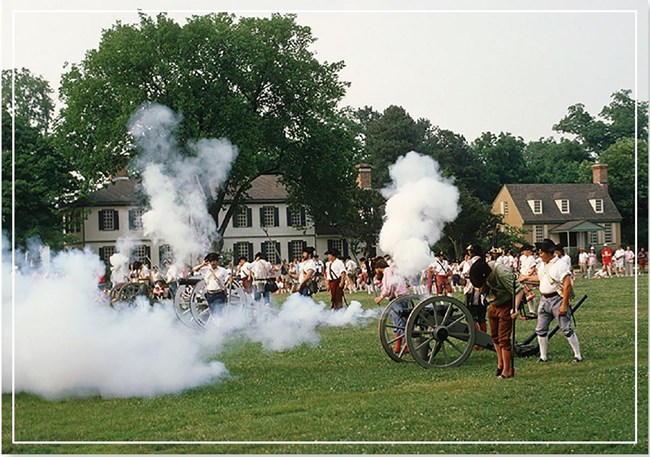 A crowd watching a cannon demonstration by historic reenactors on a grass field near historic brick buildings