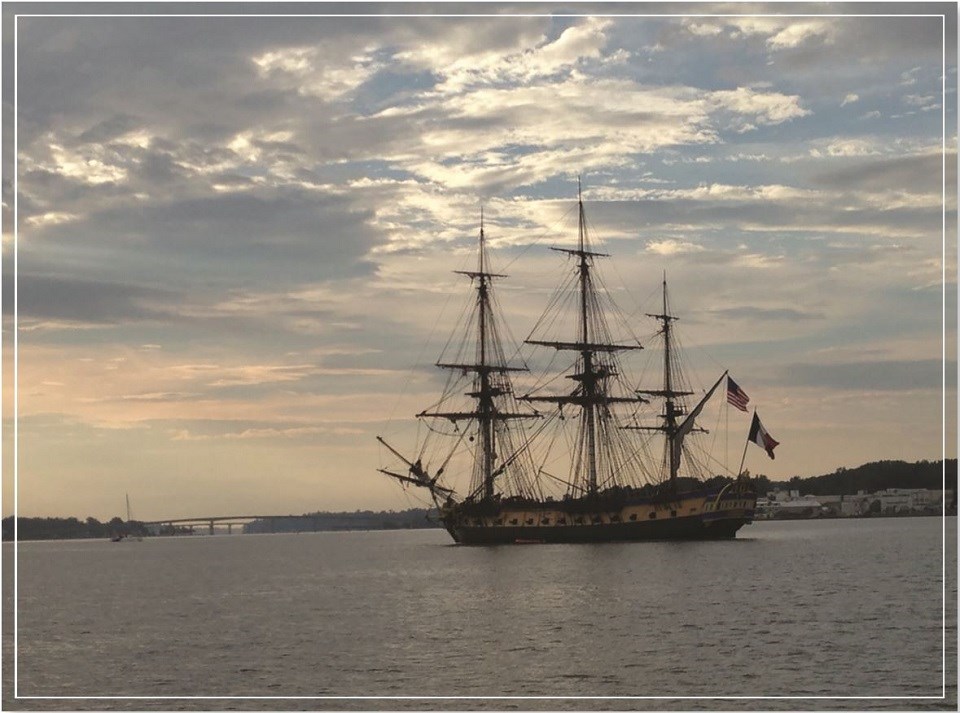 A three-masted clipper with no sails raised in a harbor. An American flag and French flag fly from its stern.