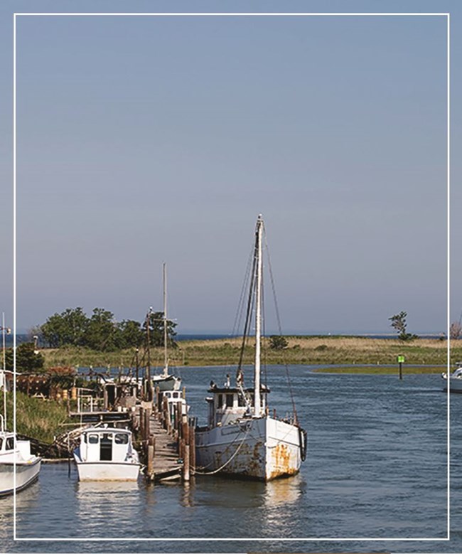 A fishing and several smaller boats are docked against a wooden pier in an inlet.