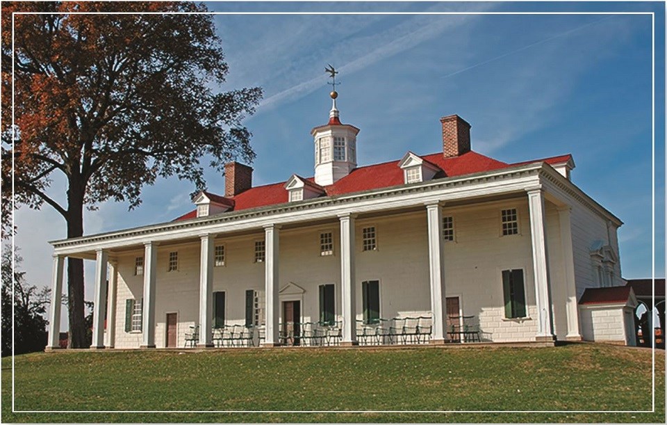 A two-story white mansion with a two-story proch supported by rectangular columns. The roof is bright red with three front-facing dormers. Two red brick chimneys flank a weathervane mounted on a lantern.