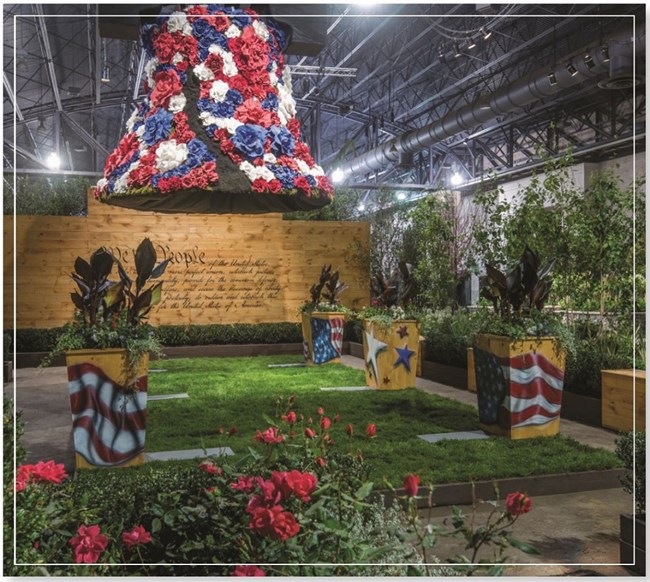 A liberty bell replica covered in red, white, and blue flowers is mounted over a small grassy area with several planters painted with colorful patriotic designs. The preamble of the Constitution is written on a wall as a backdrop.