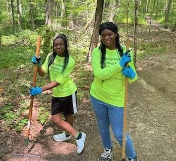 Two girls stand in a walking path in a forest with two rakes.