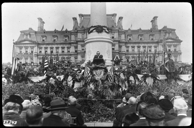 A man speaks from a lectern flanked by uniformed soldiers, flags, and bunting.