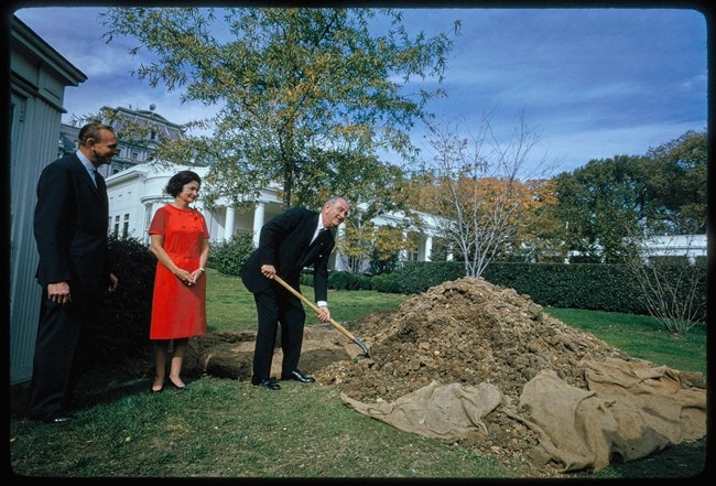 Lyndon Johnson smiles and sticks a shovel in a pile of dirt at the White House while Lady Bird Johnson and Irvin Williams look on.