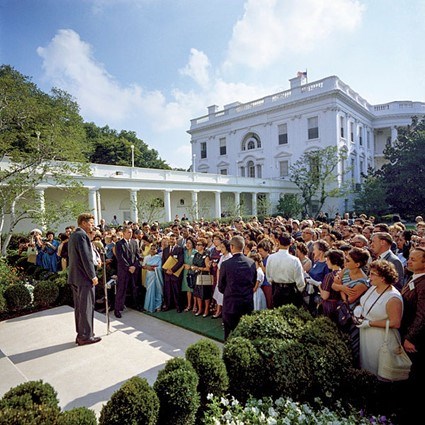 John F. Kennedy speaks before a crowd of people in the Rose Garden on a sunny day.