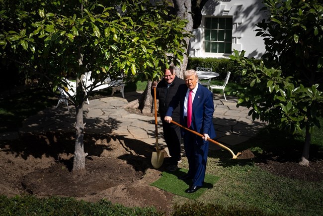 President Trump in a blue suit holds a shovel with dirt near the White House. Dale Haney stands next to him, leaning on a shovel.