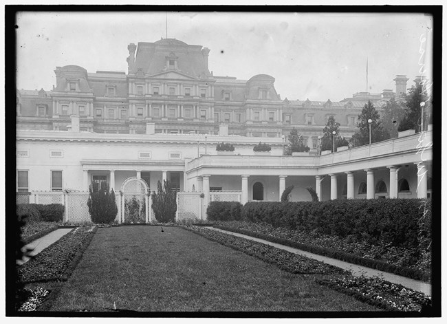 Black and White photo showing the White House rose garden designed by George E. Burnap, with view of the State, War, and Navy Building in the background.