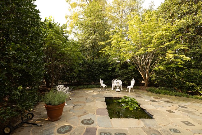 A patio with pavers and white garden chairs walled in with greenery.