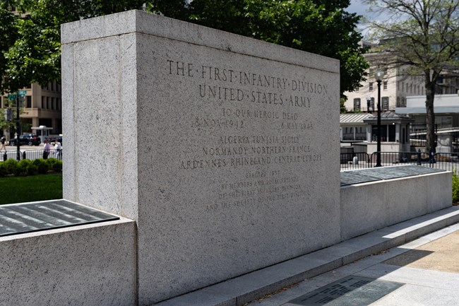 A rectangular stone monument flanked by a low, angled stone wall with bronze plaques on top.