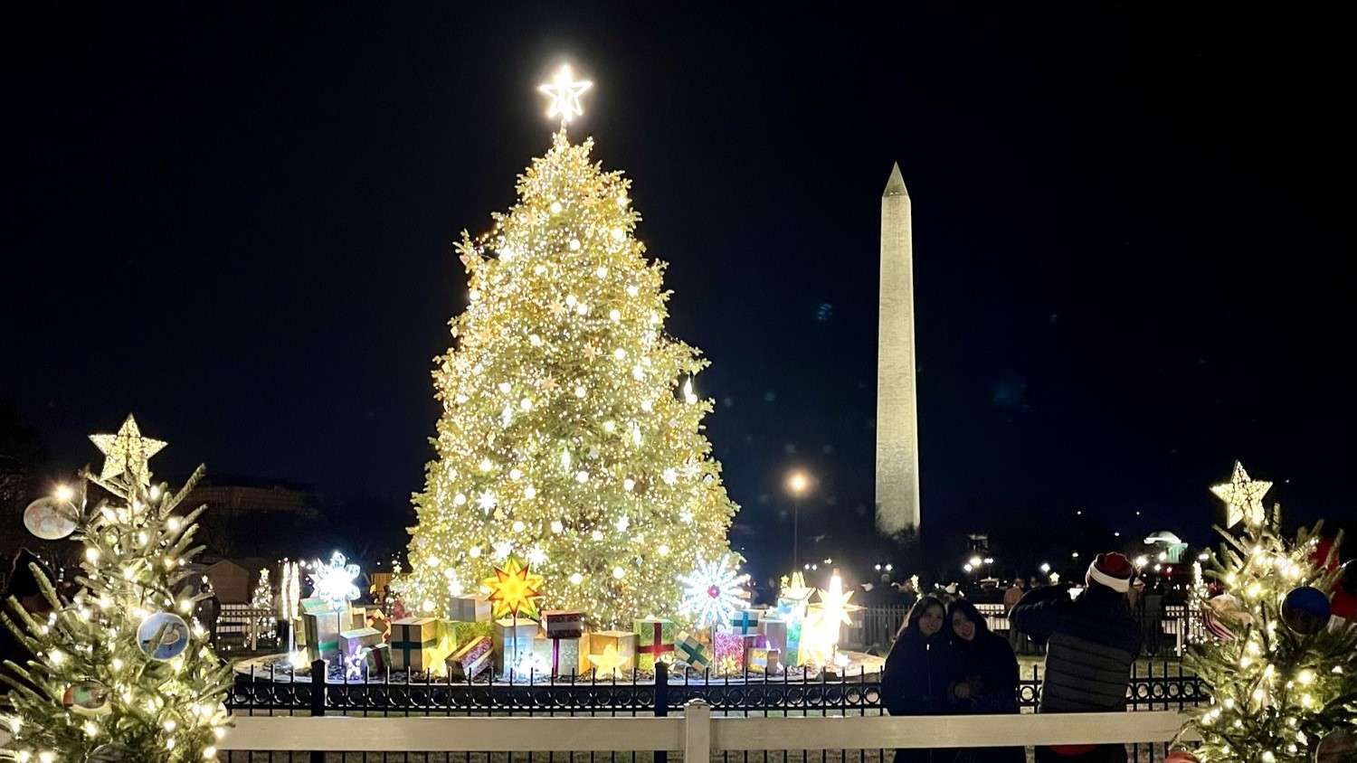 A nighttime scene with a large Christmas tree with white lights and a star on top in front of the Washington Monument.