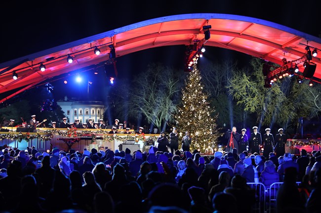 A lighted Christmas tree appears behind a stage with live performers as an audience looks on. In the background the South face of the White House is visible.