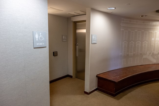 A wooden bench to the right of a doorway with a metal door to a woman's restroom.