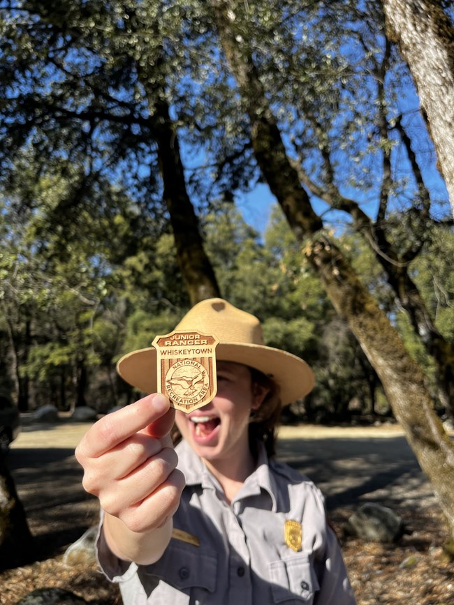 A female park ranger in flat hat and grey shirt holding up Whiskeytown's Junior Ranger badge.