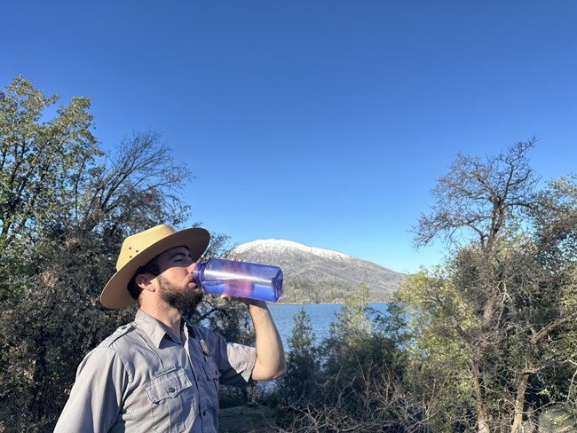 A park ranger with flat hat and grey shirt drinking water out of a reusable bottle.