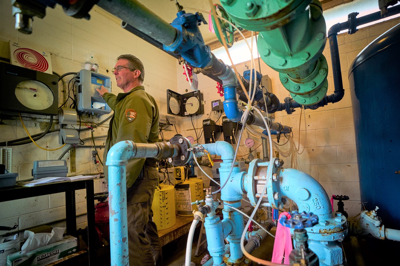 Photo of Chris Blevins inside water treatment building. Chris is wearing green jacket. Lots of pipes in background.