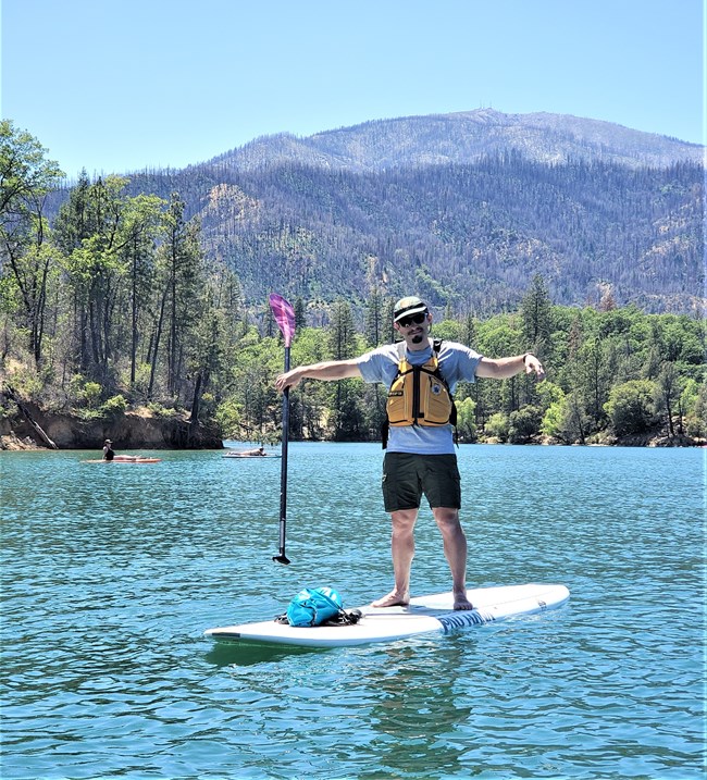 A park ranger in grey shirt, green shorts, and green ballcap standing up on paddleboard with arms and paddle out to the side. Whiskeytown Lake surrounding ranger and paddleboard. Shasta Bally (mountain) in background).