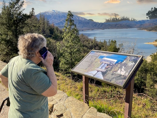 A senior citizen woman holding cell phone up towards her ear in front of Visitor Center wayside exhibit. The women is listening to the audio version of the wayside.