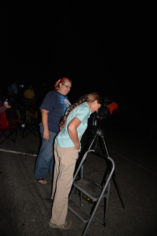At nighttime, two visitors, one looking through the lens of a telescope.