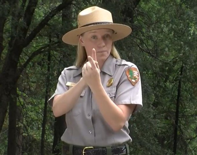 A Yosemite National Park white female park ranger in green pants, grey shirt, and flat hat, conducting American sign language.