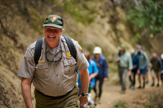 A park ranger in green pants, green ballcap, and grey shirt is all smiles while leading a hike for visitors up the trail.