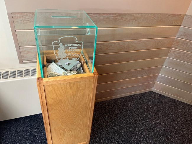 A 4 foot wooden box with a glass square top.  dollar bills are seen inside through the glass top