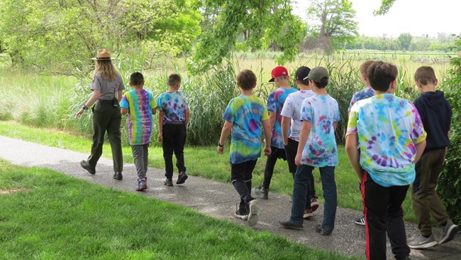 A uniformed ranger leads a group of students in tie-dye shirts down a sidewalk path