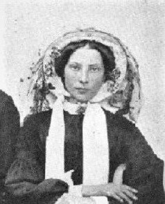A black and white photo of a young woman in black clothing and a white bonnet-like garment around her head. Her arms are cross in her lap at the bottom of the frame.