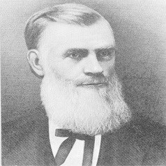 A black and white portrait from chest up of a man in a suit and bowtie with hair slicked to the side and a large white beard.