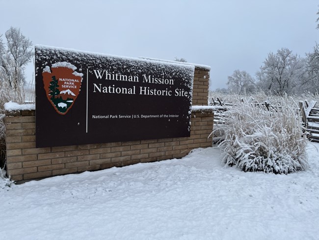 A brown sign with the text "Whitman Mission National Historic Site" on to the left of the text is the official National Park Service Arrowhead logo.  A field of snow surrounds the sign with bare deciduous trees in the background.