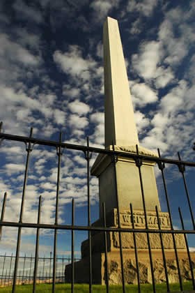 An obelisk surrounded by a picket fence with a blue sky and clouds in the background