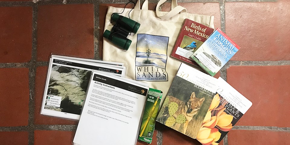 A bag surrounded by a collection of items, including books, binoculars, and laminated maps and instructions.
