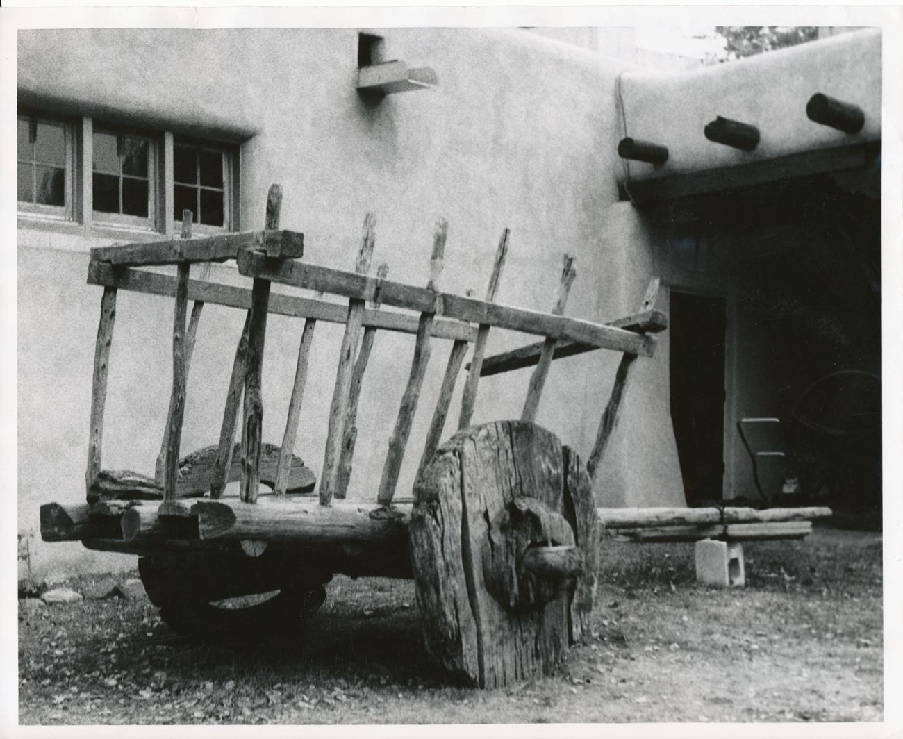 an old wooden cart with two wheels and long handles sits in front of an adobe wall of the visitor center