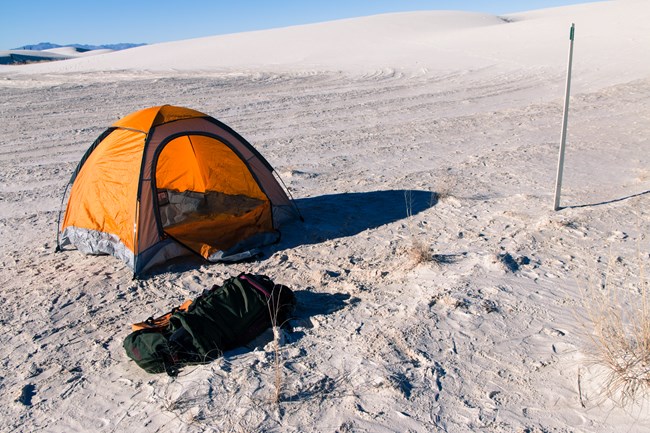 orange camping tent on dunes next to green stake in the ground. Backpacking bag on the ground in front.