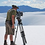 Man with camera on tripod filming dunes.