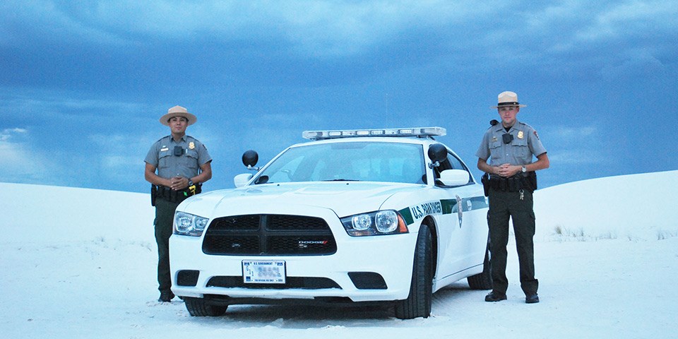 Two law enforcement rangers standing on each side of a patrol car surrounded by white sand dunes