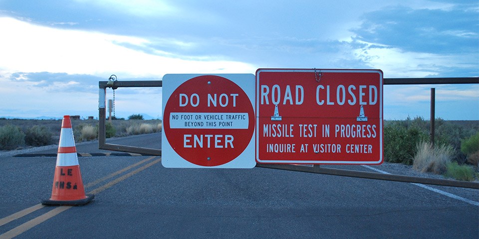 Cone and gate across road with red and white  signs reading “Do Not Enter” and “Road Closed”