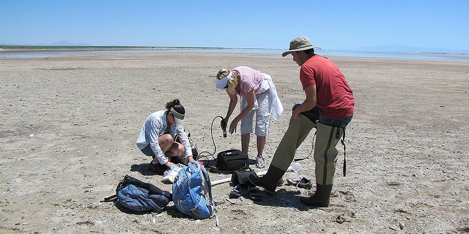 Two women and a man with waders and two blue backpacks on the flat shores of a shallow lake.