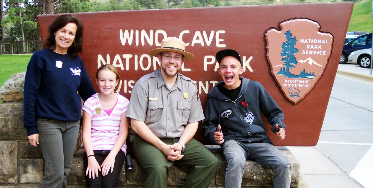 Junior rangers sitting with a park ranger in front of the Wind Cave National Park historic sign
