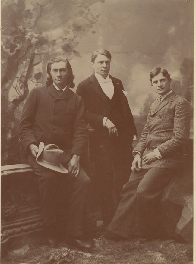 three men in a sepia photo wearing old fashioned suits posing in a studio against a backdrop