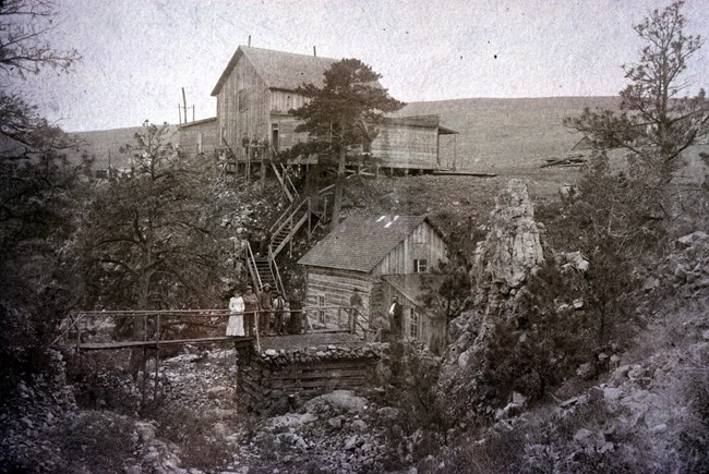 black and white photo of two old wooden buildings along a ravine, a small bridge crossing the ravine has a group of visitors in old clothing standing on it