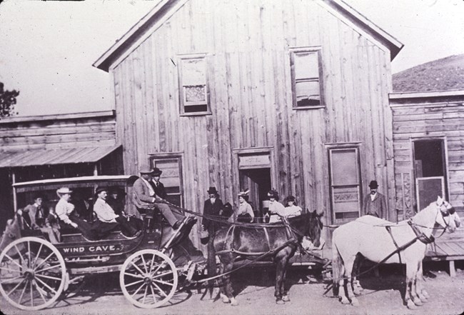 black and white photo of a stagecoach with Wind Cave written on it drawn by four horses in front of a wooden building