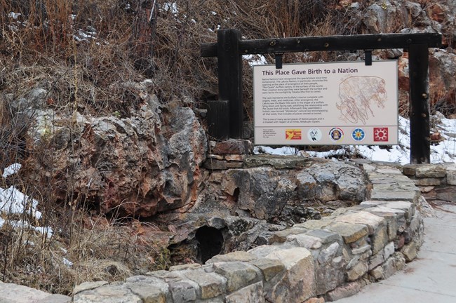 View of the natural entrance to Wind Cave in the lower left with a stone wall in the foreground and a sign describing the cultural significance of the cave to indigenous people in the background.