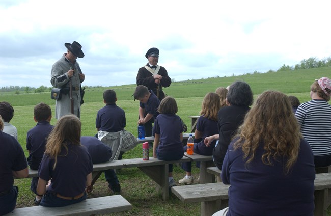 Two men in historical uniforms give a program to a group of children outside.