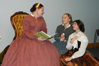 Three ladies dressed in Victorian period clothing, two are on the floor looking up at a woman sitting in a chair reading a book.