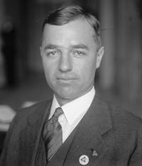 A man in a suit and tie and small white pin on his lapel stares into the camera